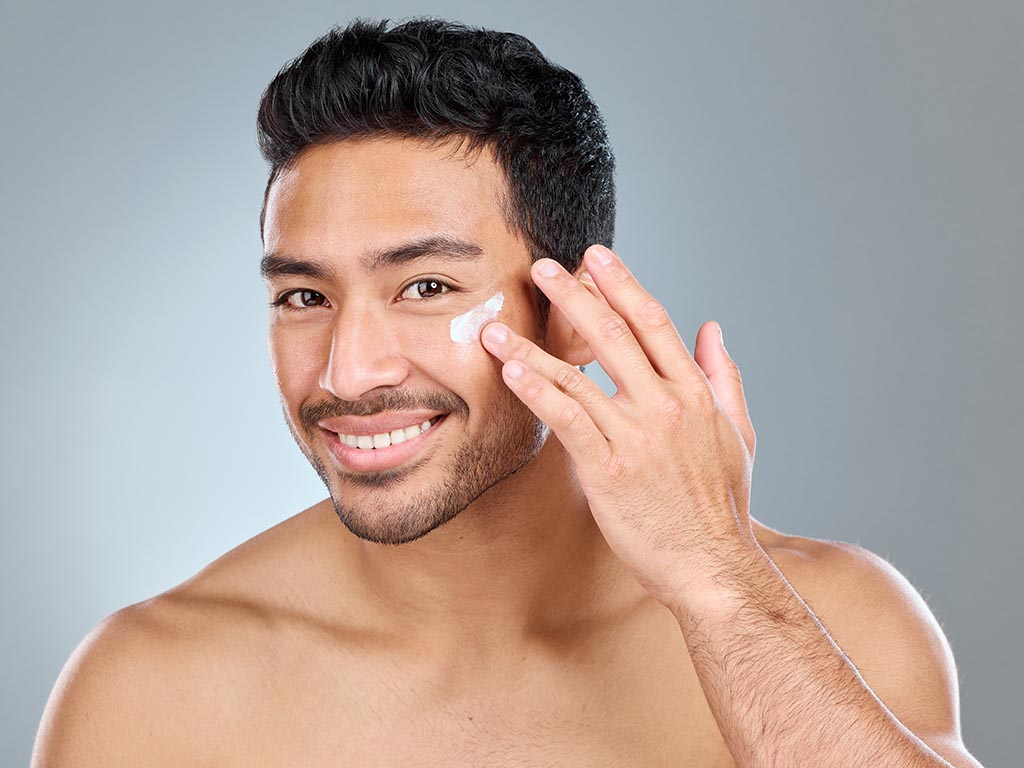 Shot of a man applying moisturiser to his face while standing against a grey background.